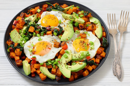 fried Sweet potato hash browns served with sunny-side-up eggs and fresh avocado slices on a plate on white wooden table with forks, close-up, not AI-generatedの写真素材