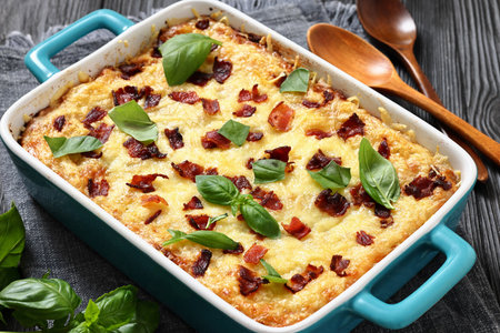 twice baked potato casserole with fried bacon and cheddar cheese in a baking dish on black wood table with spoons and basil, horizontal view, close-up, not AI generatedの写真素材