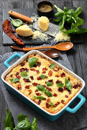 twice baked potato casserole with fried bacon and cheddar cheese in a baking dish on black wood table with spoons and ingredients, vertical view, not AI generatedの写真素材