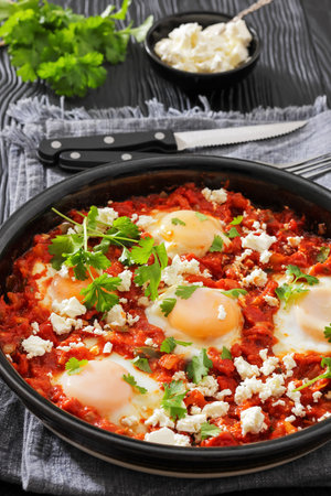 shakshuka, eggs poached in rich tomato vegetable sauce sprinkled with crumbled feta cheese and cilantro in a baking dish on wood table with cutlery, vertical view, close-up, not AI generatedの写真素材