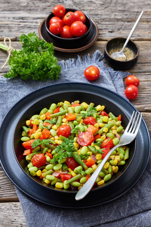 succotash, american dish of cooked corn, edamame beans, tomatoes, green beans in black bowl on rustic wood table with fork and parsley, vertical view, not AI generatedの写真素材