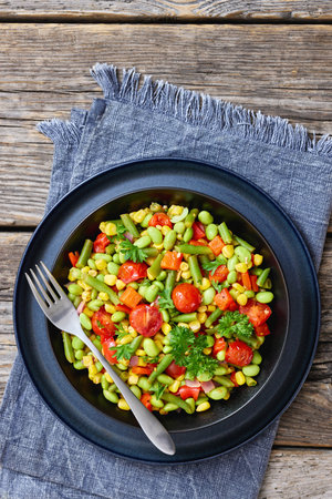 succotash, classic American side dish of cooked corn, edamame beans, tomatoes, green beans in black bowl on rustic wood table with fork , flat lay, vertical view, not AI generatedの写真素材