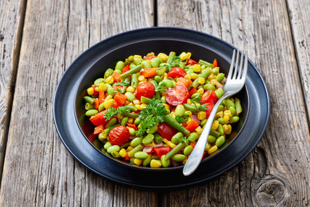 succotash, classic American side dish of cooked corn, edamame beans, tomatoes, green beans in black bowl on rustic wood table with fork, horizontal view, not AI generatedの写真素材
