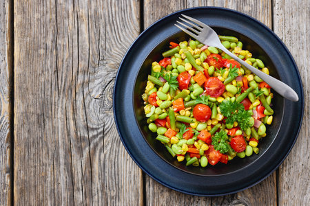 succotash, classic American side dish of cooked corn, edamame beans, tomatoes, green beans in black bowl on rustic wood table with fork, flat lay, free space, not AI generatedの写真素材