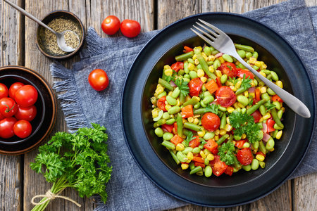 succotash, classic American side dish of cooked corn, edamame beans, tomatoes, green beans in black bowl on rustic wood table with fork and ingredients, flat lay, not AI generated, close-upの写真素材