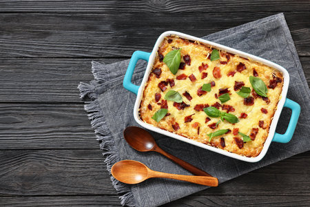 twice baked potato casserole with fried bacon and cheddar cheese in a baking dish on black wood table with spoons and napkin, horizontal view, flat lay, free space, not AI generatedの写真素材