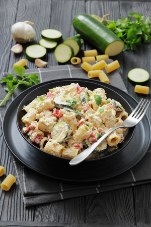 chicken alfredo rigatoni with zucchini and red bell pepper in silky creamy sauce in a black bowl on wood table with ingredients, vertical view from above,  not AI generatedの写真素材
