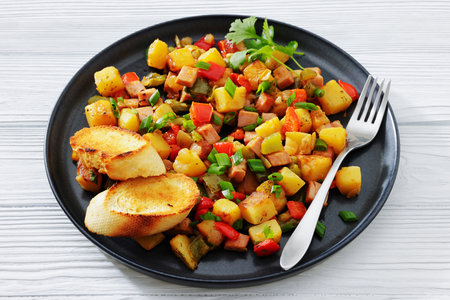 crispy fried cubed potatoes with ham, bell peppers and onion sprinkled with green onion on a plate with toasts and fork on white wood table, horizontal view,  close-upの写真素材