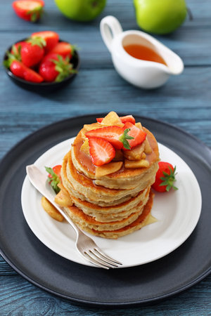 stack of pancakes topped with caramelized apple slices and fresh strawberries poured with maple sirup on plate with dessert fork on wood table, vertical view, not AI generatedの写真素材