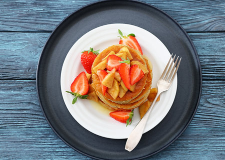 stack of pancakes topped with caramelized apple slices and fresh strawberries poured with maple sirup on plate with dessert fork on rustic wood table, flat lay, close-up, not AI generatedの写真素材