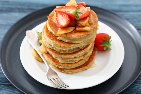 stack of pancakes topped with caramelized apple slices and fresh strawberries poured with maple sirup on plate with dessert fork, horizontal view, not AI generatedの写真素材