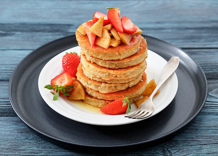 delicious pancakes topped with caramelized apple slices and fresh strawberries poured with maple sirup on plate with dessert fork, close-up, not AI generatedの写真素材