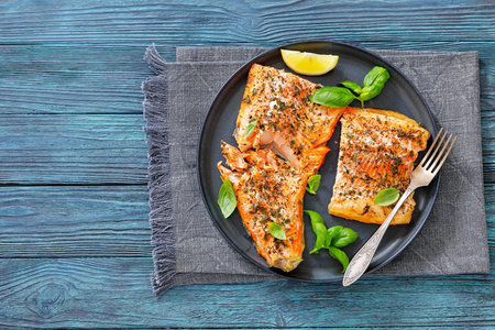 grilled whole salmon fillets with skin-on on a plate with fresh basil, lemon slices, fork on wood...