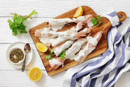 Greek style raw chicken souvlaki marinade with yogurt and seasoning on wood sticks on cutting board on white wood table with kitchen towel, cilantro and lemon, flat lay, not AI generatedの写真素材