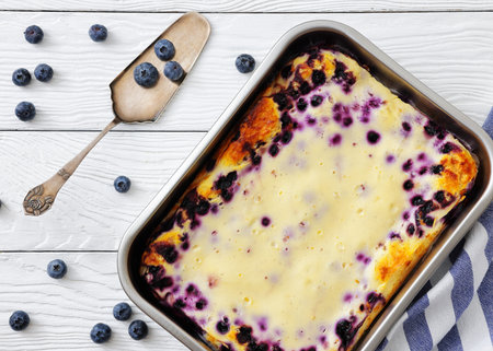 Freshly baked Mustikkapiirakka, Finnish Blueberry pie in a baking dish on white wood table with cake shovel and tea towel, close-up, flat lay, horizontal view from above, not AI generatedの写真素材