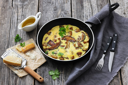 porcini mushroom parmesan omelette on a skillet on rustic wood table with cutlery, gratified cheese and olive oil, horizontal view from above, not AI generatedの写真素材
