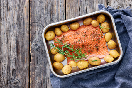 raw salmon fillet with baby potatoes and fresh rosemary in baking dish prepared for bake in oven on rustic wood table, horizontal view from above, flat lay, free space, not AI generatedの写真素材