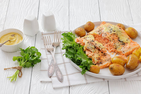 Salmon fillet topped with melted cheese, spices, and baby potatoes on a white plate with fork on white wood table with forks and mustard, horizontal view, close-up, not AI generatedの写真素材