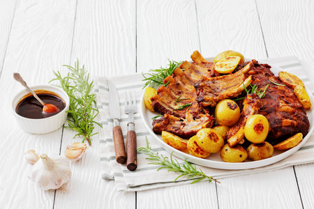 bbq baby pork ribs with potatoes, fresh rosemary on a plate on white wood table with cutlery, sauce, garlic, horizontal view from above, not AI generatedの写真素材