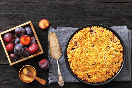 Crumb topped Plum Pie in a baking dish on dark wood table with cake shovel, brown sugar and fresh plums, horizontal view from above, flat lay, free space, not AI generatedの写真素材