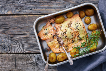 Salmon fillet topped with melted cheese, spices, rosemary, and baby potatoes in a baking dish on rustic wood table, horizontal view, flat lay, free space, not AI generatedの写真素材