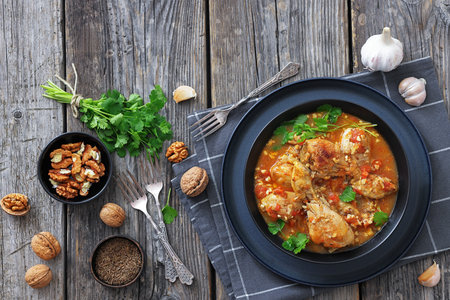 Chakhokhbili, Georgian chicken stew with tomato, herbs and chopped walnuts in a black bowl on a rustic wood table with ingredients, forks, horizontal view, flat lay, free space, not AI generatedの写真素材