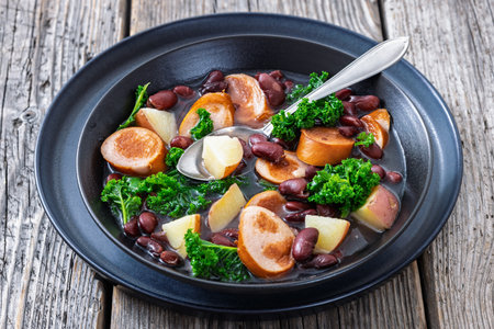 Red kidney bean, potato, kale and sausage soup in a black bowl with spoon on rustic wood table, horizontal view from above, not AI generatedの写真素材