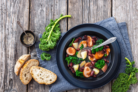 Red kidney bean, potato, kale and sausage soup in a black bowl with spoon on rustic wood table with bread, horizontal view from above, flat lay, free space, not AI generatedの写真素材