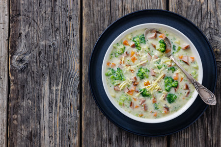 Sausage Vegetable Chowder, thick cheesy soup with chunks of broccoli, carrot, potato in white bowl with spoon, on rustic wood table, flat lay, free space, horizontal view from aboveの写真素材