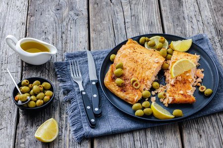 roasted salmon fillet on plate with green olives and lemon slices on rustic wood table with cutlery, horizontal view from above, not AI generatedの写真素材