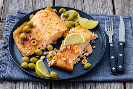 baked salmon fillet on grey plate with green olives and lemon slices on rustic wood table with cutlery, horizontal view from above, not AI generatedの写真素材