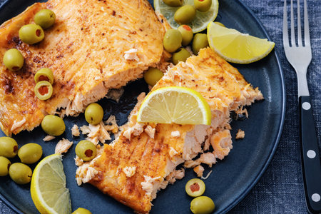 roasted salmon fillet on plate with green olives and lemon slices on rustic wood table with cutlery,...