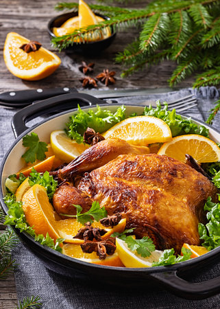 Whole roasted crispy golden skin chicken in a baking dish with sliced oranges and lettuce on table with cutlery, fir branches, pine cones, vertical view, close-up, not AI generatedの写真素材