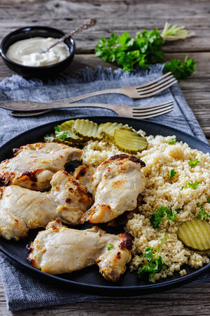 Grilled boneless chicken thighs served with couscous, pickles on white plate on a rustic wooden table with Greek yogurt dip and forks, vertical view, close-up, not AI generatedの写真素材