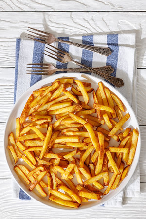 crispy golden french fries on a plate with napkin and forks on white wooden table, vertical view,...