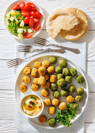 Green and golden falafel balls served with creamy hummus dip, pita bread, and a fresh cucumber tomato salad on a light wooden table, vertical, not AI generated