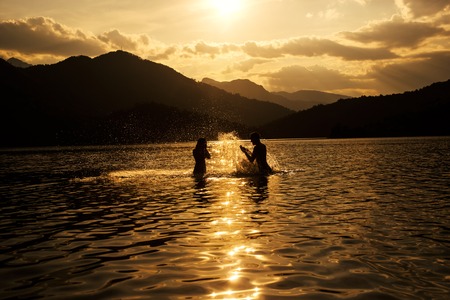 Silhoueted young couple at the beach on a hazy summer day at dusk,  enjoying splashing in the ocean water, getting wet,の写真素材