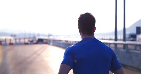 Attractive fit man running fast along big modern bridge at sunset light, black man doing workout outdoors, silhouette runner in windbreaker jogging over bridge road with amazing sunset on backgroundの写真素材