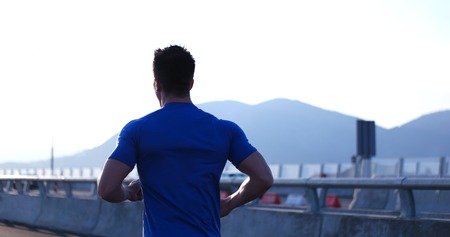 Attractive fit man running fast along big modern bridge at sunset light, black man doing workout outdoors, silhouette runner in windbreaker jogging over bridge road with amazing sunset on backgroundの写真素材
