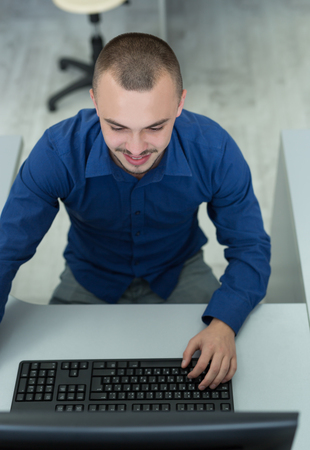 Handsome young businessman working at his desktop computer sitting at his desk in the officeの写真素材