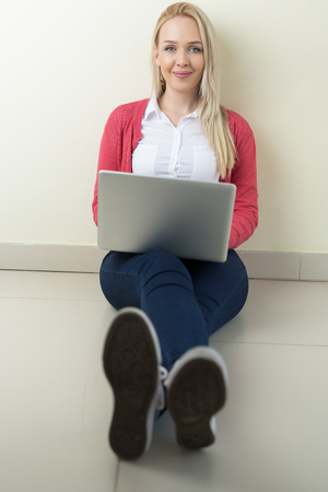 Happy young woman sitting on the floor with crossed legs and using laptopの写真素材