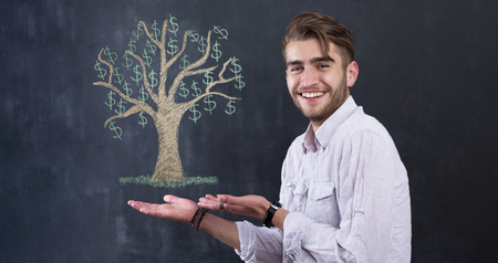 Young teenager man holding a tree with dollars in a front of chalkboard background. Growing business conceptの写真素材