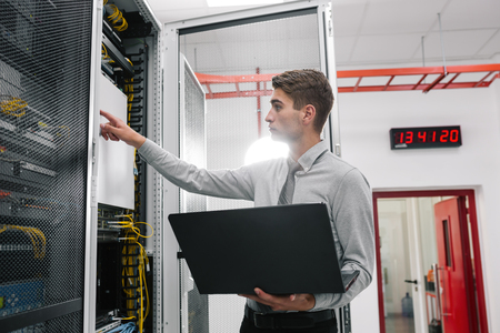 Portrait of modern young man holding laptop standing in server room working with supercomputerの写真素材