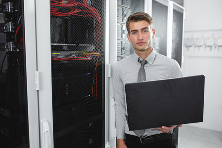 Portrait of modern young man holding laptop standing in server room working with supercomputerの写真素材