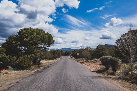 Empty road between trees under blue sky and clouds in Hill near Santa Fe, New Mexico, USAの写真素材