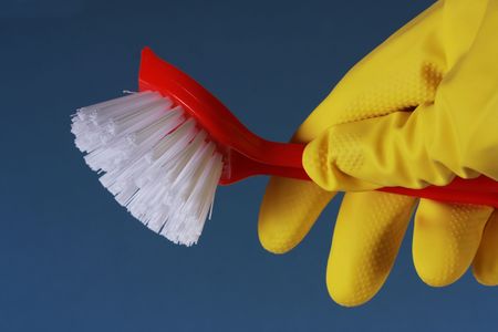 Closeup of a yellow rubber glove holding a red washing up brush against a blue background.