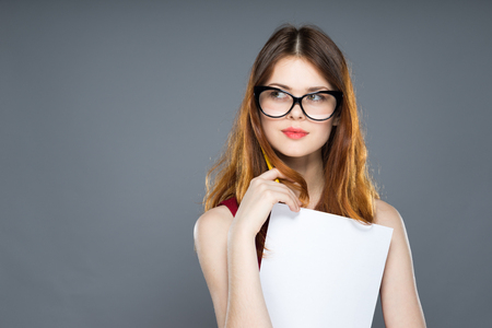 Schoolgirl in glasses keeps a sheet of paper over gray background isolated, holding white A4 paper poster, holds a pen, thoughtful faceの写真素材