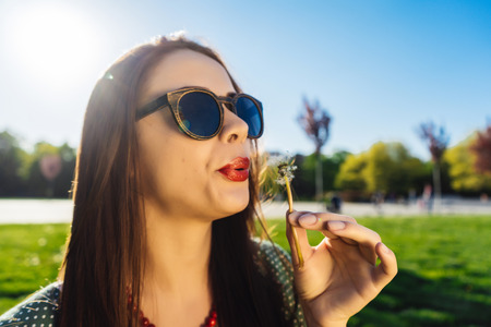 A happy smiling woman in dark sunglasses in the park sits on the lawn and blows on a dandelion, holds a dandelion in her hands. Looks to the rightの写真素材
