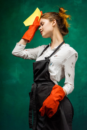 Tired woman cleaning woman in a black apron, wearing orange gloves wipes sweat from her faceの写真素材