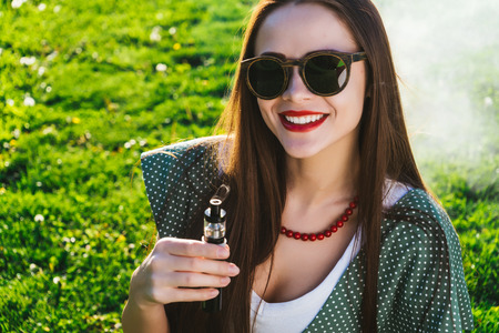 Beautiful woman with long hair sitting on the lawn with bright green grass, smokes, smiles, looks at...
